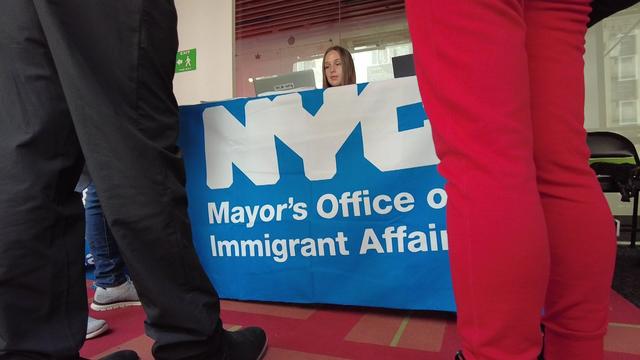 A woman sits behind a desk with a tablecloth that says NYC Mayor's Office of Immigrant Affairs. 